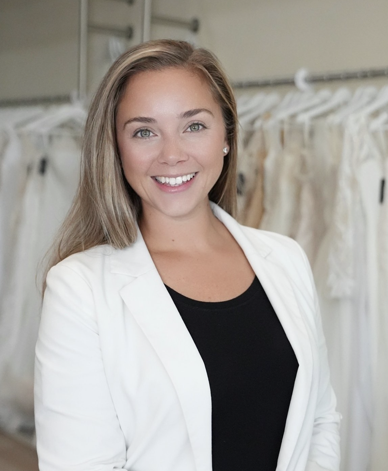 A woman with long hair wearing a white blazer and black top smiles at the camera. She stands in front of a clothing rack with white garments.