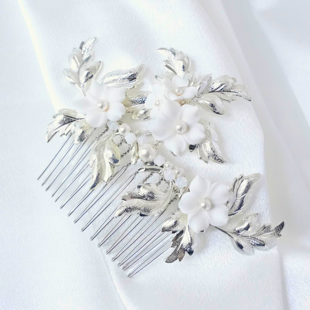 A Lavlii decorative comb with white flowers, silver leaves, and pearls rests on a white fabric—perfect for bridal accessories or special occasions.