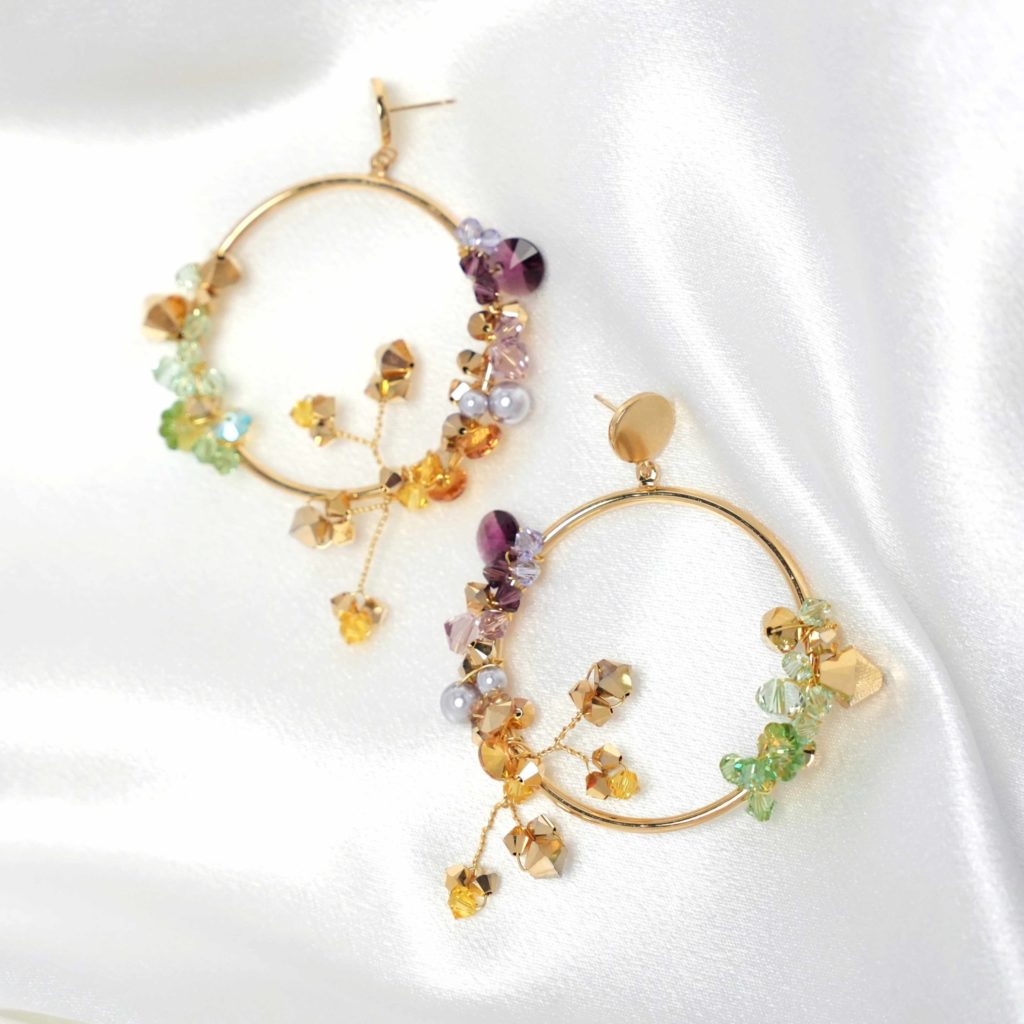A pair of Lavlii gold hoop earrings adorned with colorful beads, pearls, and crystals placed on a white satin fabric, exemplifies radiant elegance.